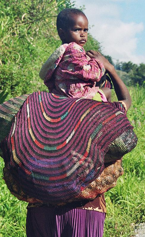 Papua1-18-Zoellner.jpg - Dani woman carrying nets with sweet potatos and her child, near Wamena, Jayawijaya regency (2008)(Photo by Siegfried Zöllner)